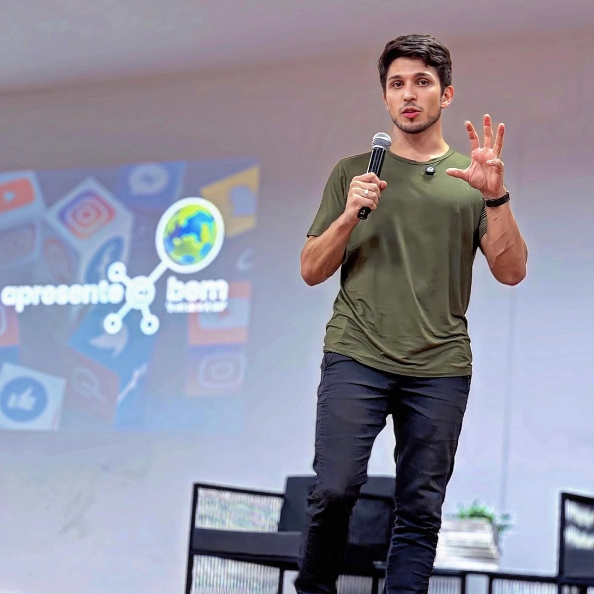 Man in olive green shirt speaking at conference with microphone, gesturing with hand raised, presentation screen visible behind